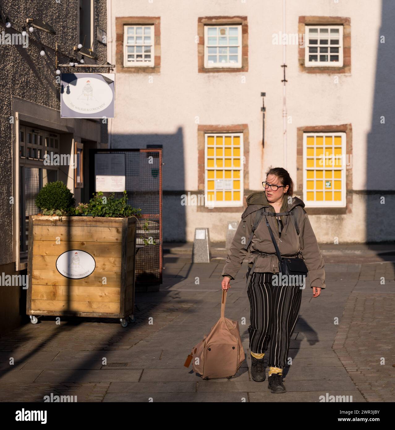 7 March 2024. Inverness,Highlands and Islands,Scotland. This is a woman pulling her luggage bag along a lane and into the sunlight in Inverness City c Stock Photo