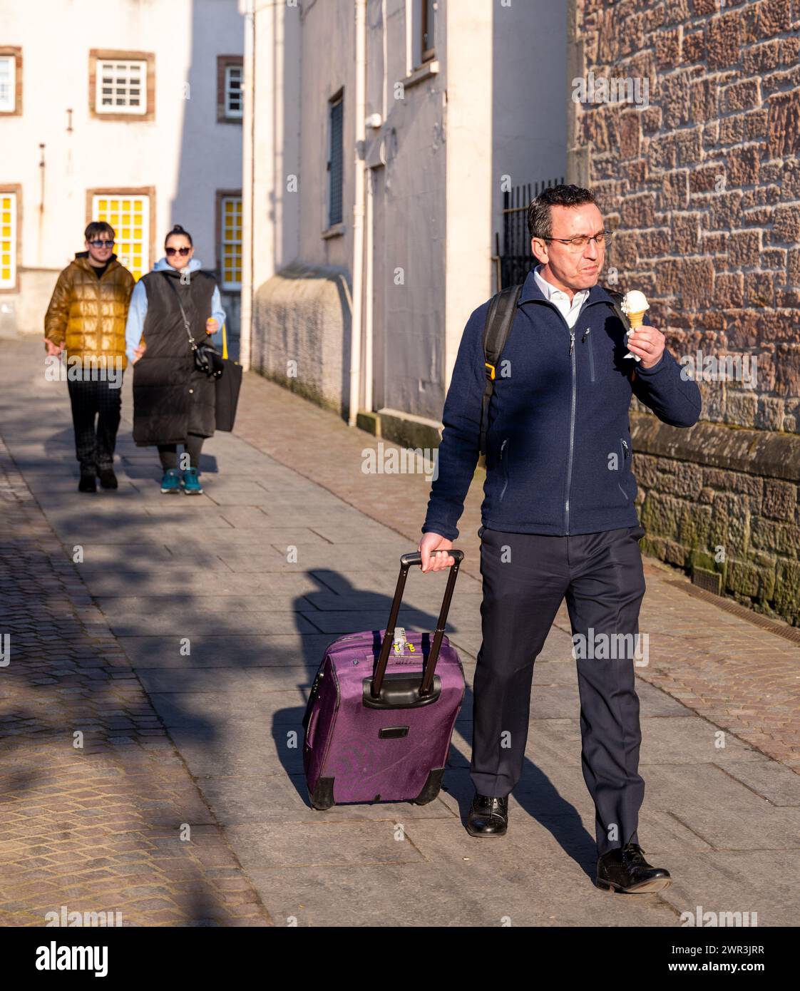 7 March 2024. Inverness,Scotland. This is a man with his luggage walking into the sunshine with his ice cream within a lane in Inverness City. Stock Photo