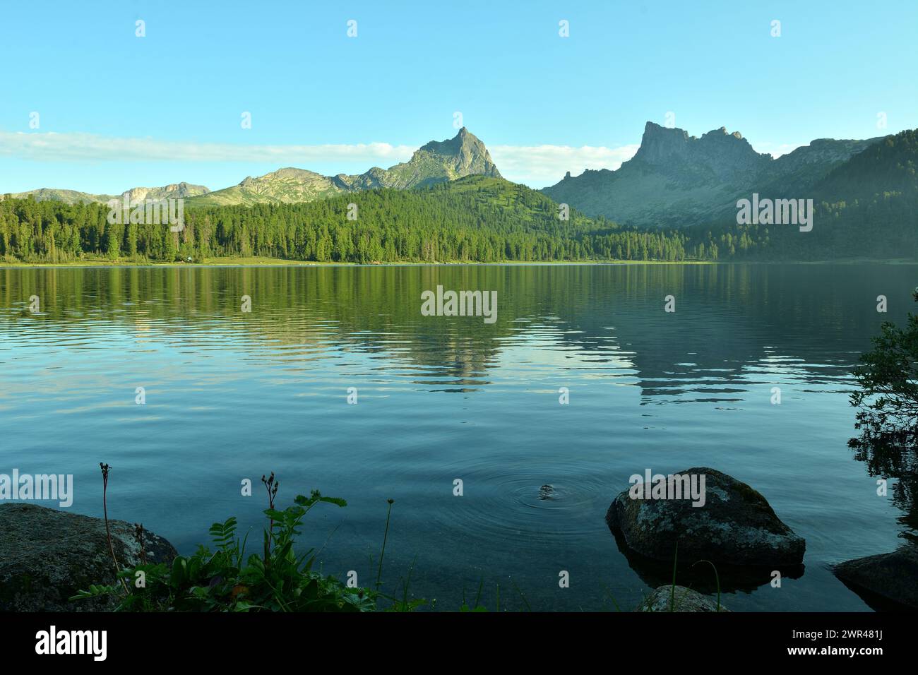 he rocky shore of a large lake in the mountains with a reflection of a dense forest and high rocks in the unsteady surface of the water on a sunny sum Stock Photo