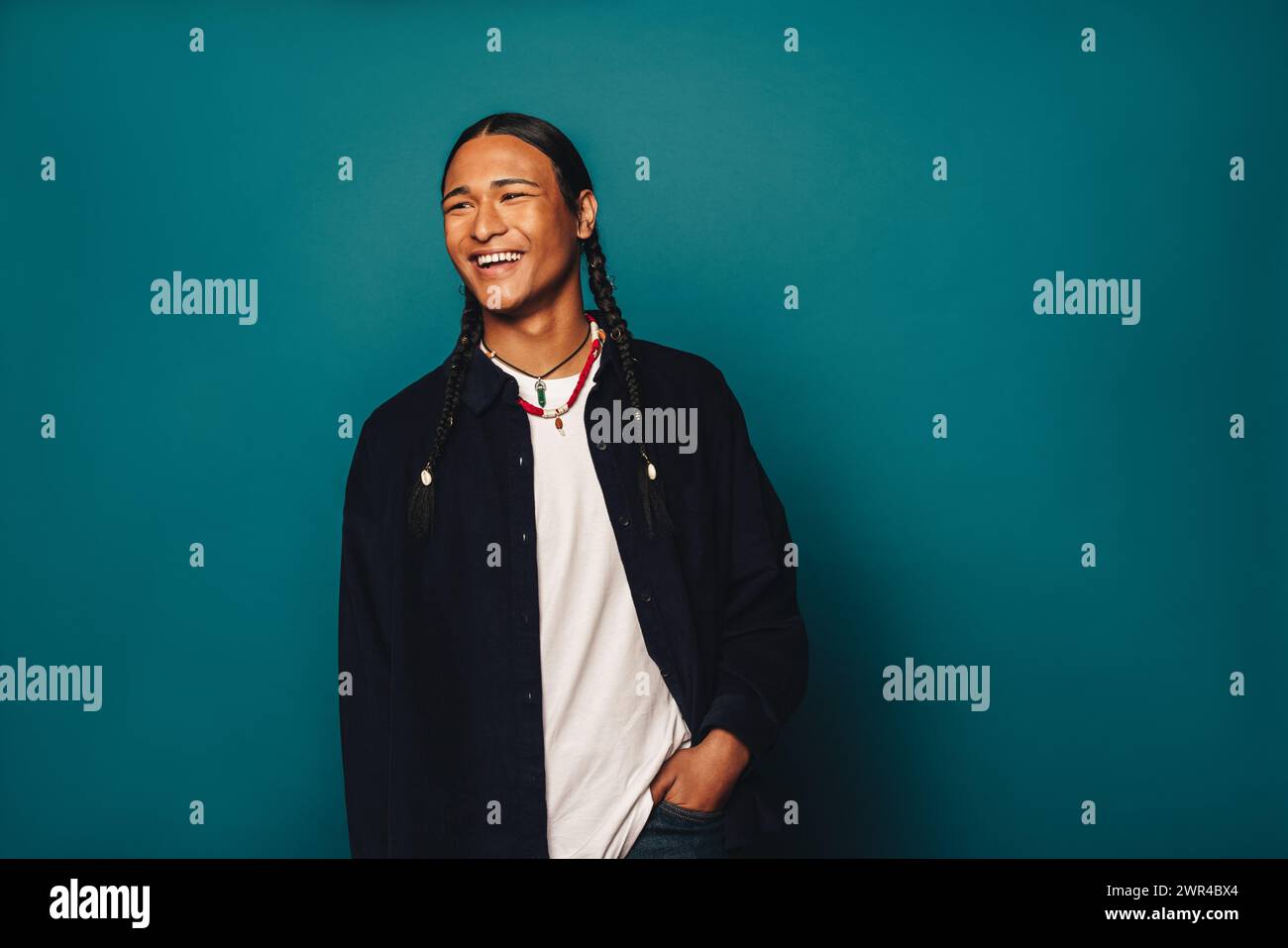 Happy young man with braided hair stands confidently against a blue background. His casual clothing and ethnic accessories add to his fashionable appe Stock Photo