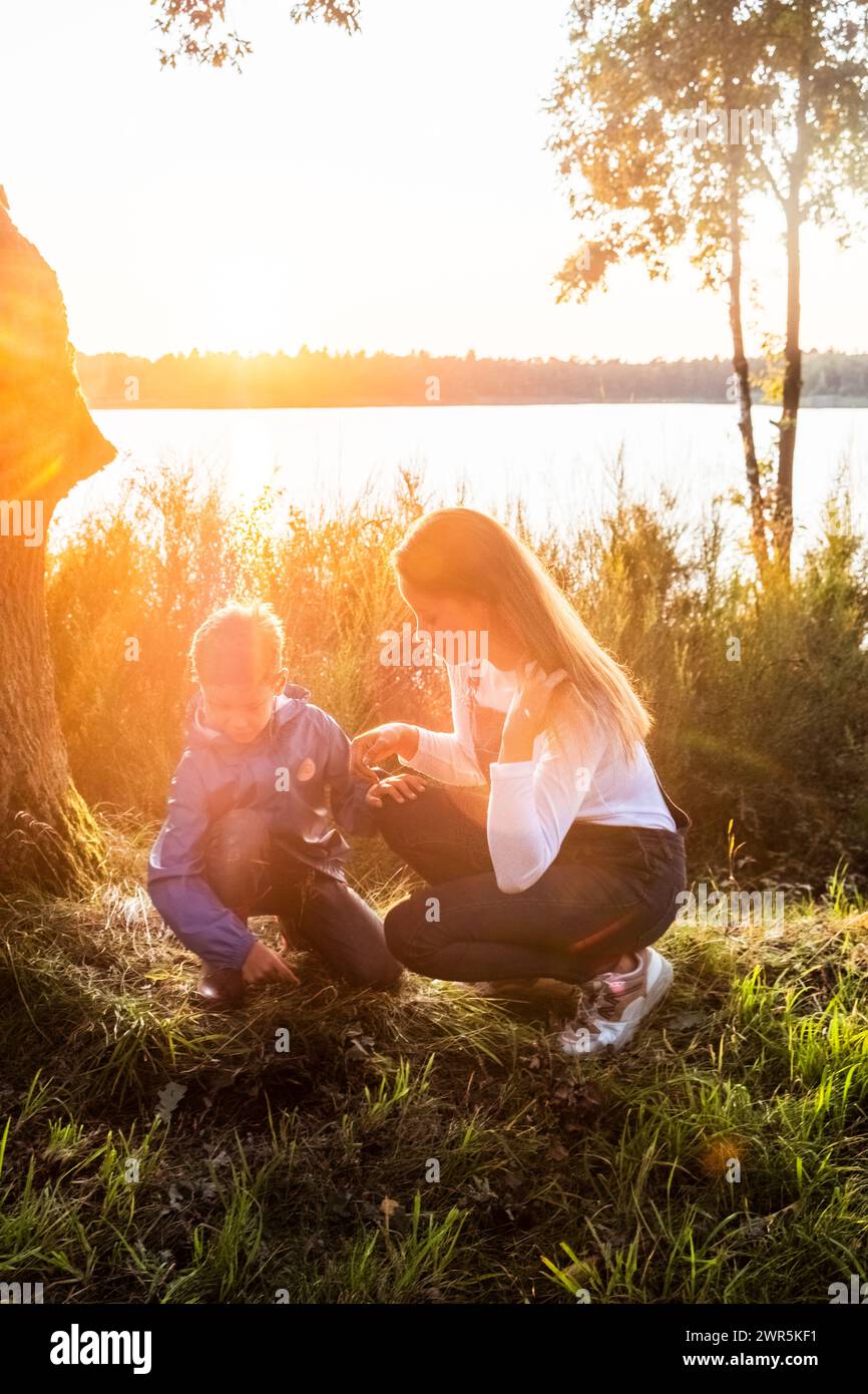 This heartwarming image captures a tender moment between a mother and her young child during a beautiful lakeside sunset. The soft, golden sunlight filters through the trees, illuminating the scene with a radiant glow. The mother, crouching down to her child's level, engages in a gentle and educational interaction, possibly exploring the nature around them. The child, dressed in a blue jacket, looks on with curiosity and wonder. The scenic backdrop of the calm lake and the surrounding foliage enhances the serene and nurturing atmosphere of the moment. Mother and Child Enjoying Lakeside Sunset  Stock Photo