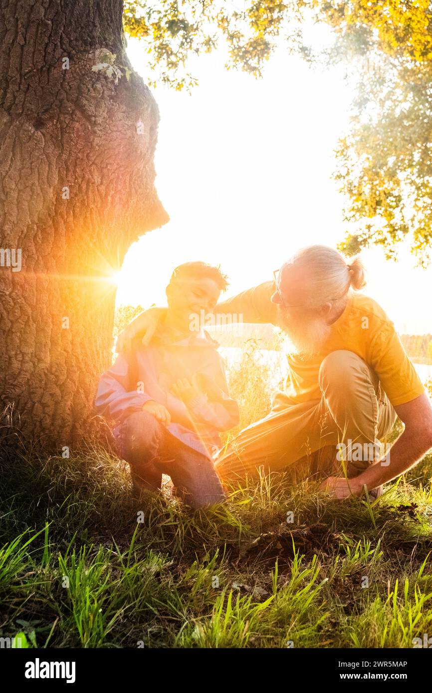This evocative image captures a heartwarming interaction between a grandfather and his grandchild in the soft, golden light of the setting sun. The older man, with a white beard and wearing a yellow shirt, is squatting down to be at eye level with the young child, who is also crouching. They are by a large tree, and the sun flares through the branches, surrounding them with a warm, luminous atmosphere. The grandfather seems to be sharing wisdom or a story with the child, who listens intently. It's a picture of generational bond and the simple, precious moments that form the fabric of family me Stock Photo