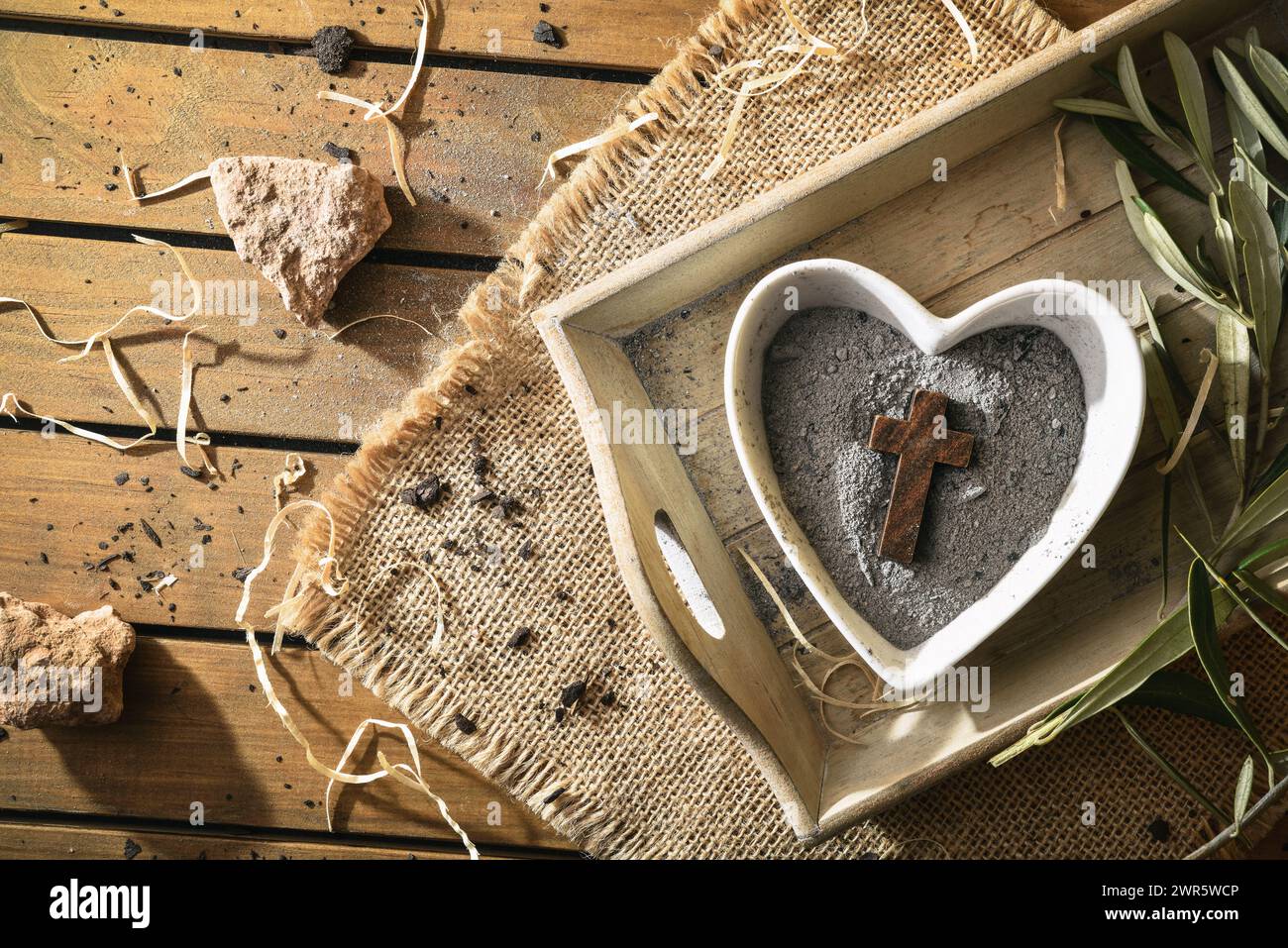 Heart-shaped container with ashes and wooden cross on wooden tray with olive leaves on a table with burlap cloth. Top view. Stock Photo