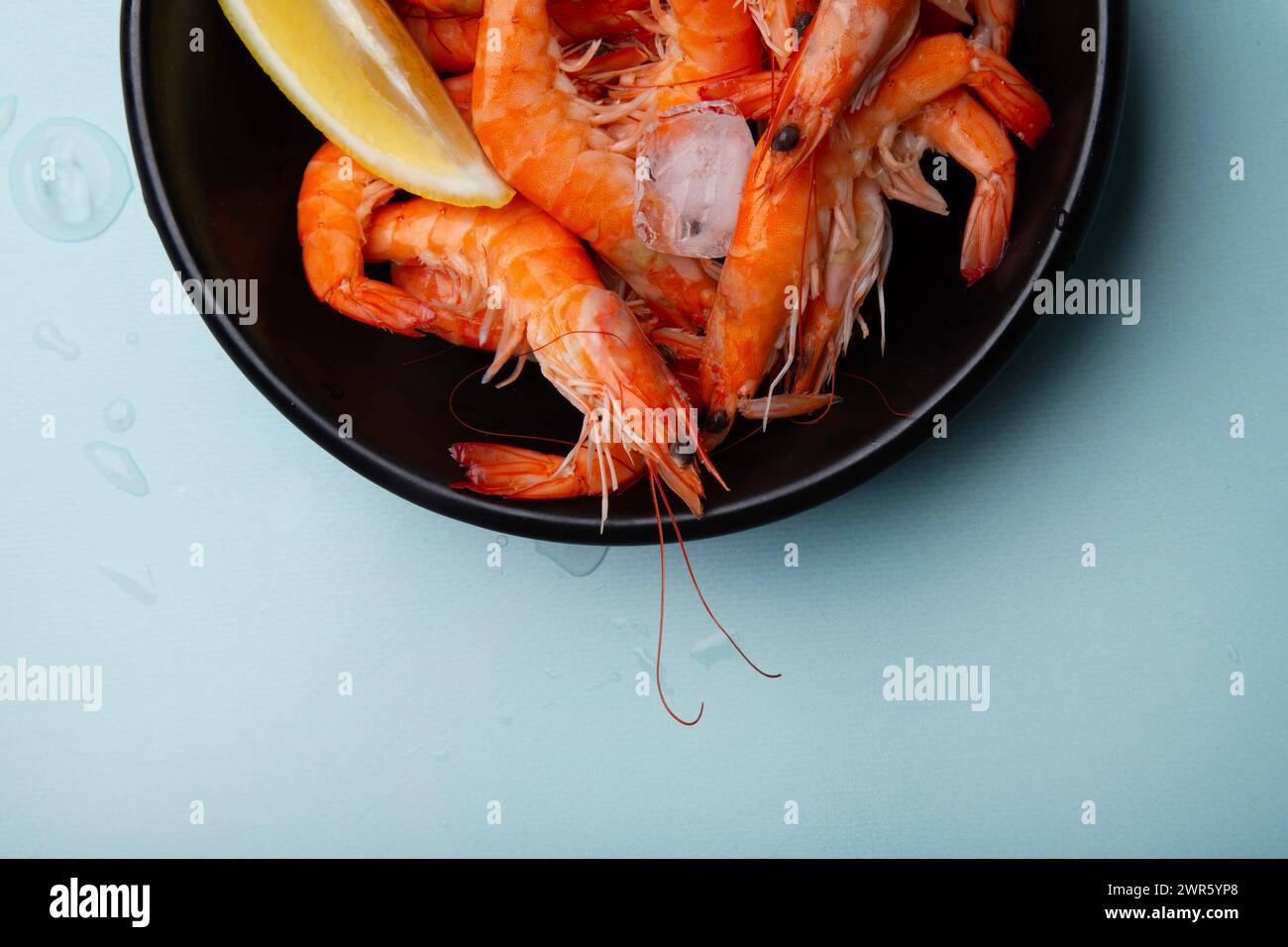 Bowl full of defrosted cooked prawns with lemon wedges, on a blue background with droplets of water. Ideal for culinary websites or seafood recipes. Stock Photo