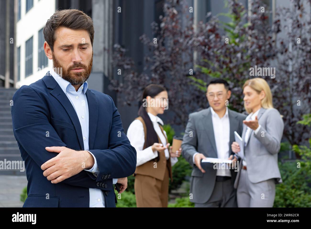 Bullying at work. Upset man standing outside office center with hands folded and head bowed, women and men team laughing at him and discussing behind his back. Stock Photo