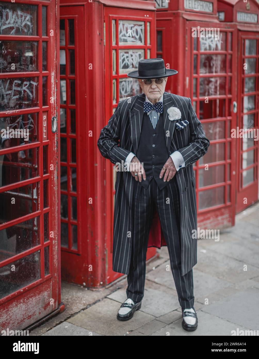 A stylish octogenarian man in a pinstripe suit poses by a row of red telephone boxes in London. Stock Photo