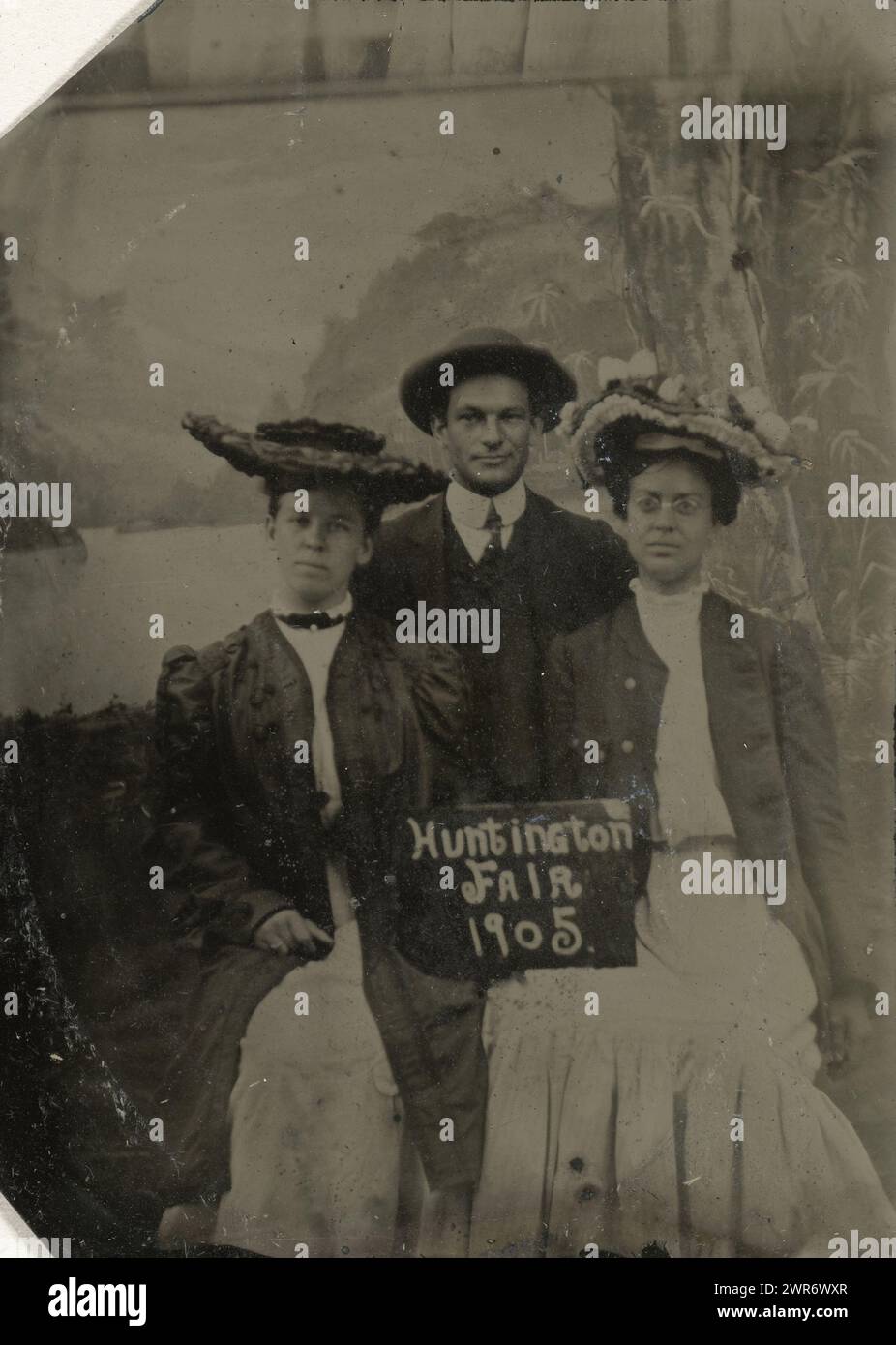 Portrait of a man and two women in front of a painted background canvas (tree, lake and mountains) and a sign with the text 'Huntington FAIR 1905', in passepartout, anonymous, United States of America, 1905, metal, ferrotyping, height 74 mm × width 51 mm, photograph Stock Photo