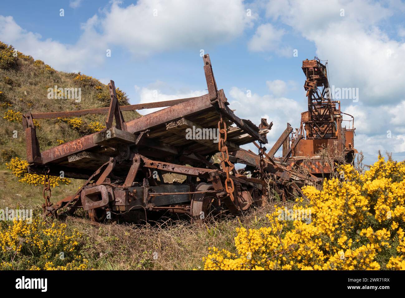 Rusty old derelict train railway crane with heavy chains wheels lifting crane on Alderney Channel Islands with yellow gorse and blue sky Stock Photo