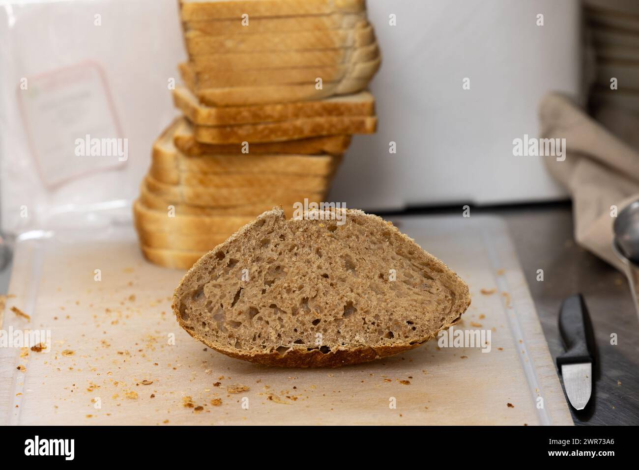 The image captures a half-slice of whole grain bread resting on a wooden cutting board with crumbs around it, suggesting the slicing process has just occurred. In the background, stacked slices of white bread are slightly blurred, contrasting with the whole grain piece in the foreground. A knife lies to the side, indicating the bread was freshly cut, likely in a home or bakery kitchen. Sliced Whole Grain Bread on Cutting Board. High quality photo Stock Photo