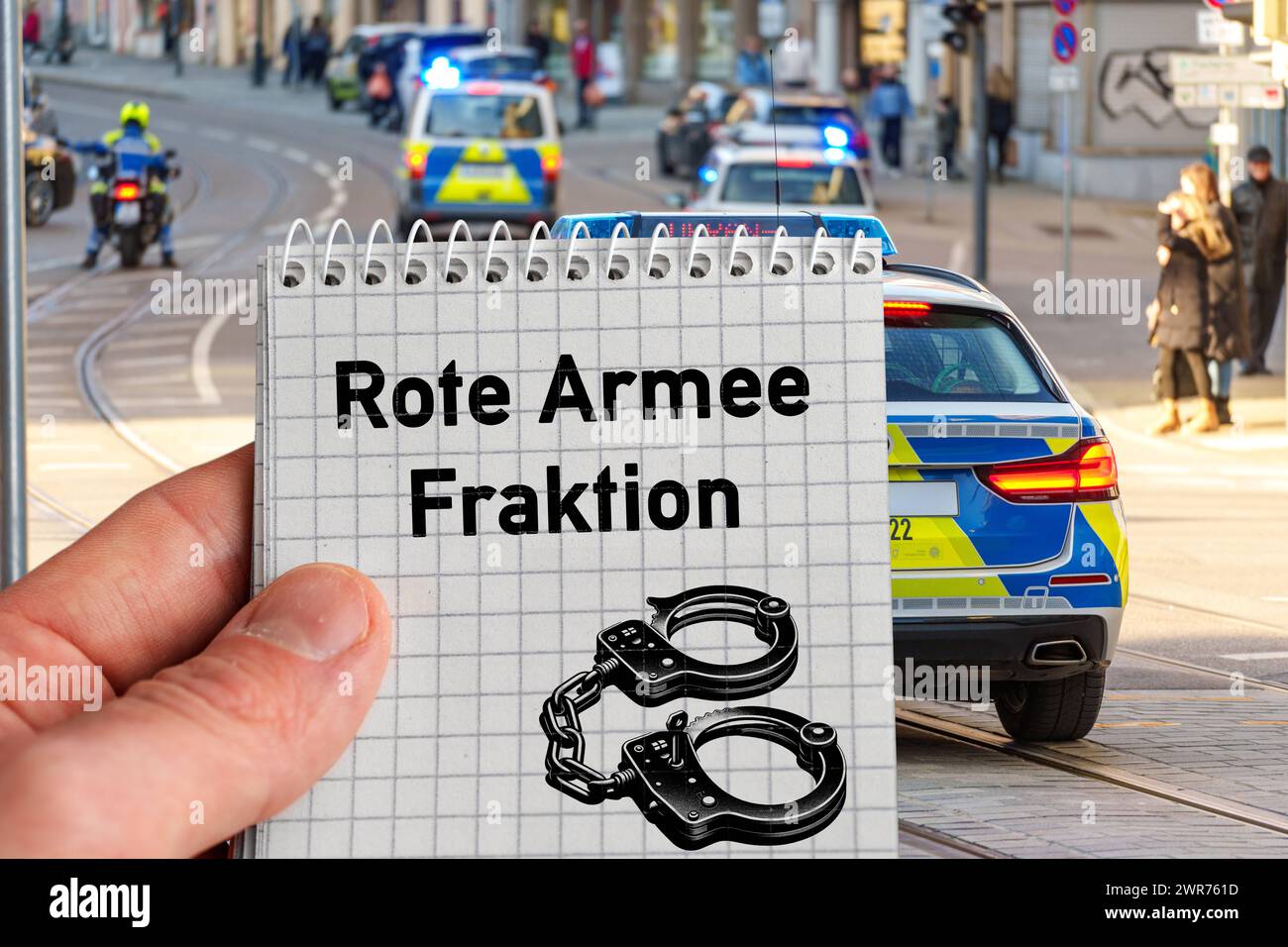 Germany - March 11, 2024: Hand holding block with inscription: Red Army Faction and handcuffs symbol in front of police emergency vehicles. PHOTOMONTAGE *** Hand hält Block mit Aufschrift: Rote Armee Fraktion und Handschellen Symbol vor Polizei Einsatzfahrzeuge. FOTOMONTAGE Stock Photo