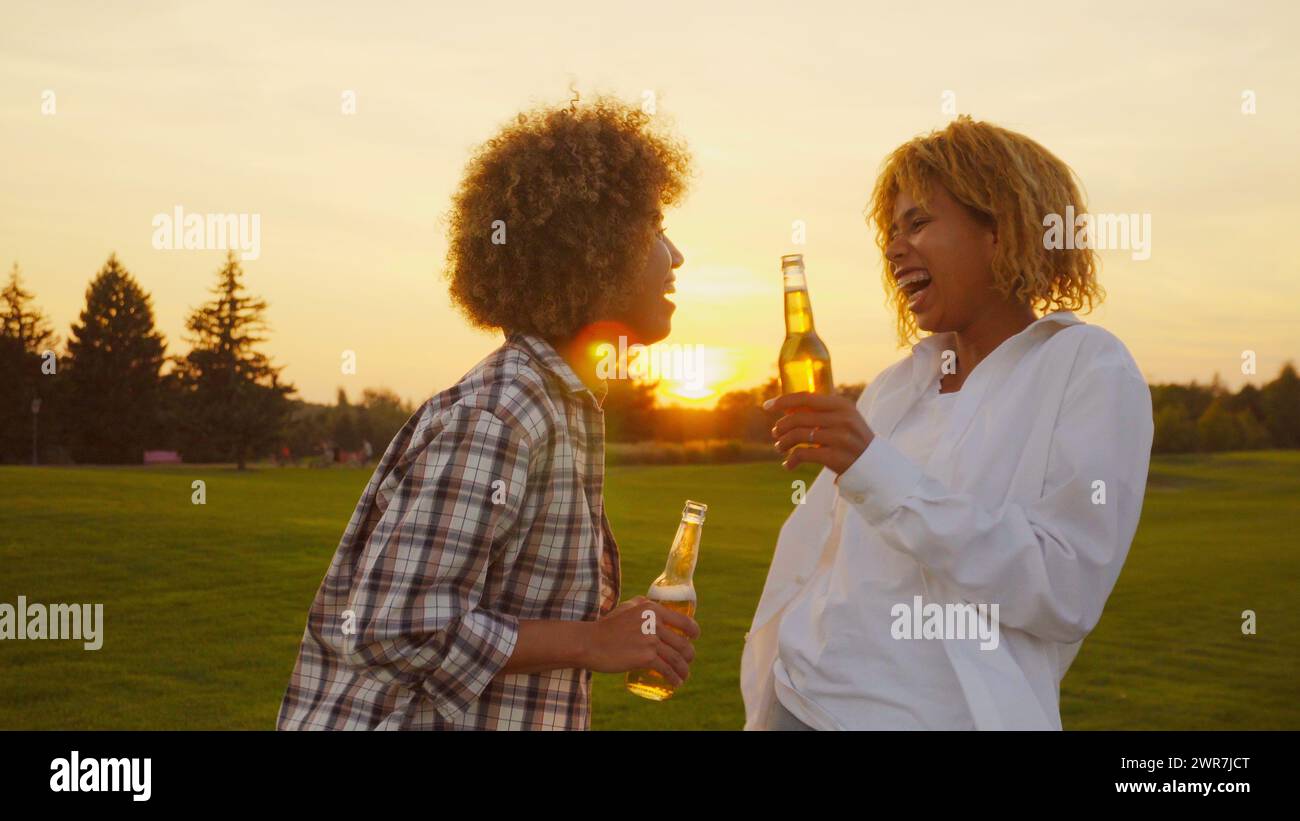 Happy multiracial young Multi-ethnic African girlfriends with afro hair having fun and drinking some beers outdoors in summertime at golden sunset Stock Photo