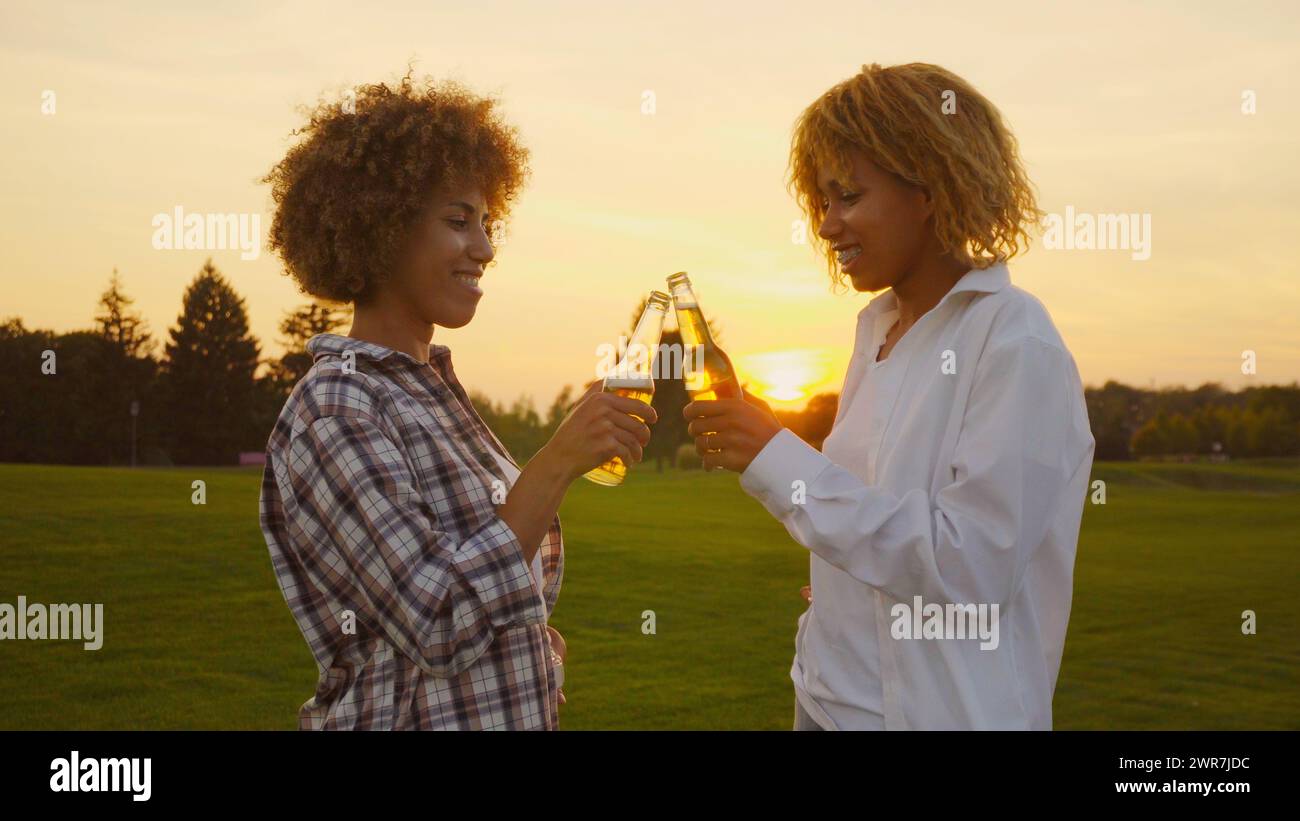 Happy multiracial young Multi-ethnic African girlfriends with afro hair having fun and drinking some beers outdoors in summertime at golden sunset Stock Photo