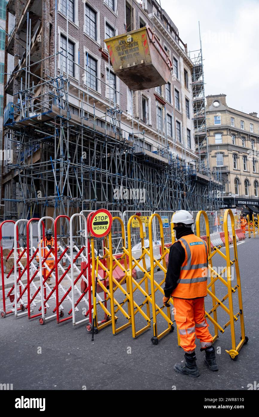 Construction worker attends barriers while he watches a skip being hoisted into the air by crane on a major construction site in Covent Garden on 6th March 2024 in London, United Kingdom. Stock Photo