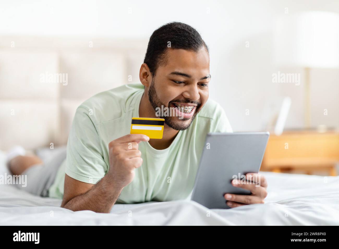 Black guy lying on bed, using digital tablet, bank card Stock Photo