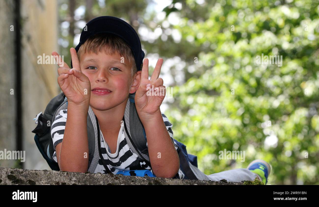 Portrait of a blond boy in a cap in the forest doing the victory gesture with both hands Stock Photo