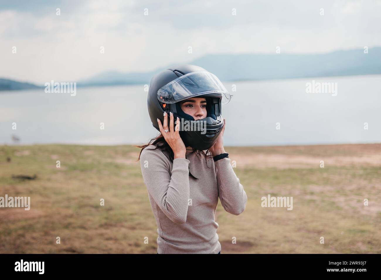 A female motorcyclist fastens her helmet against a serene lake backdrop Stock Photo