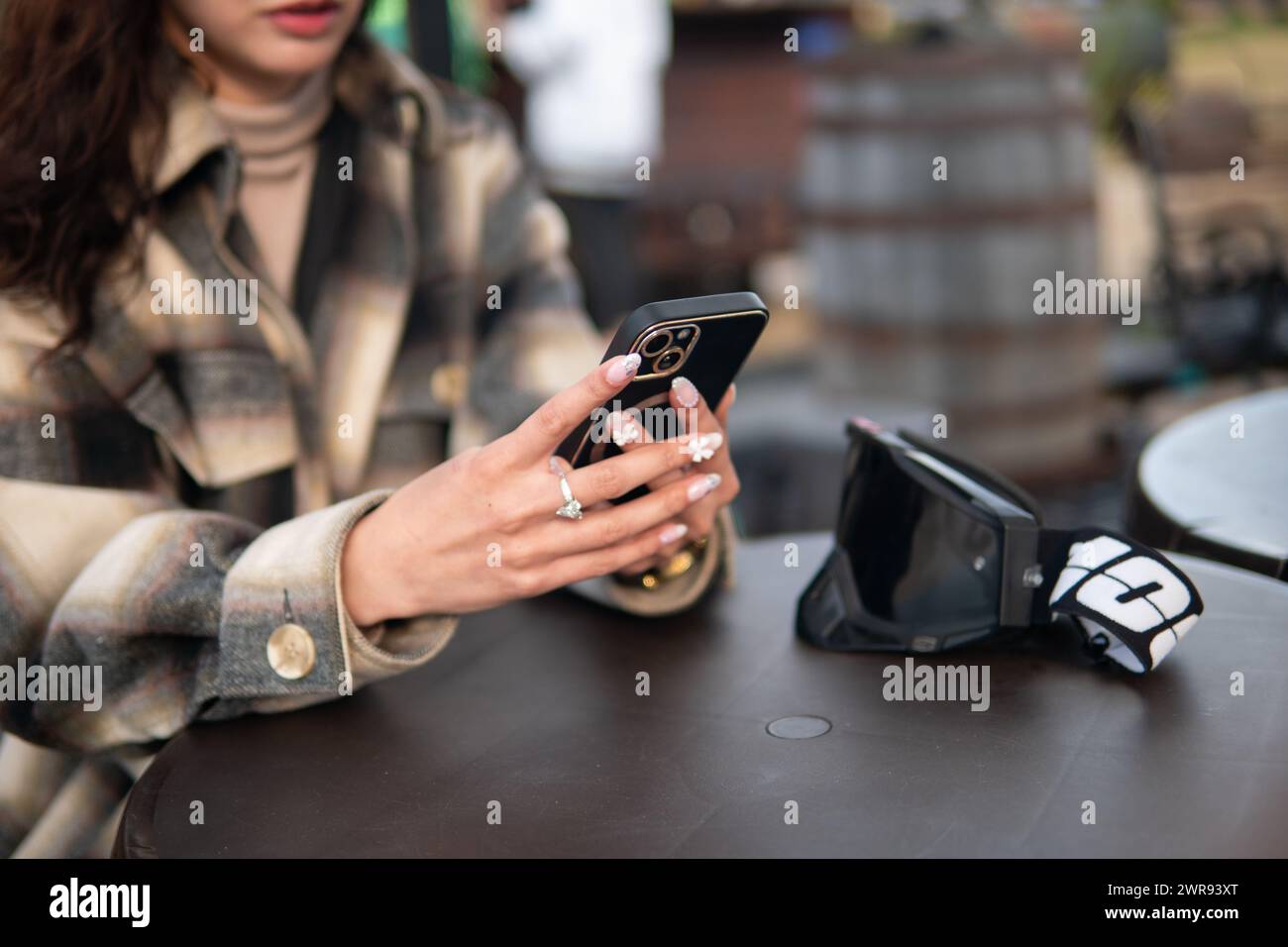 Close-up of a Latina woman's hands holding a smartphone, with motocross goggles on the table, at an outdoor cafe setting Stock Photo
