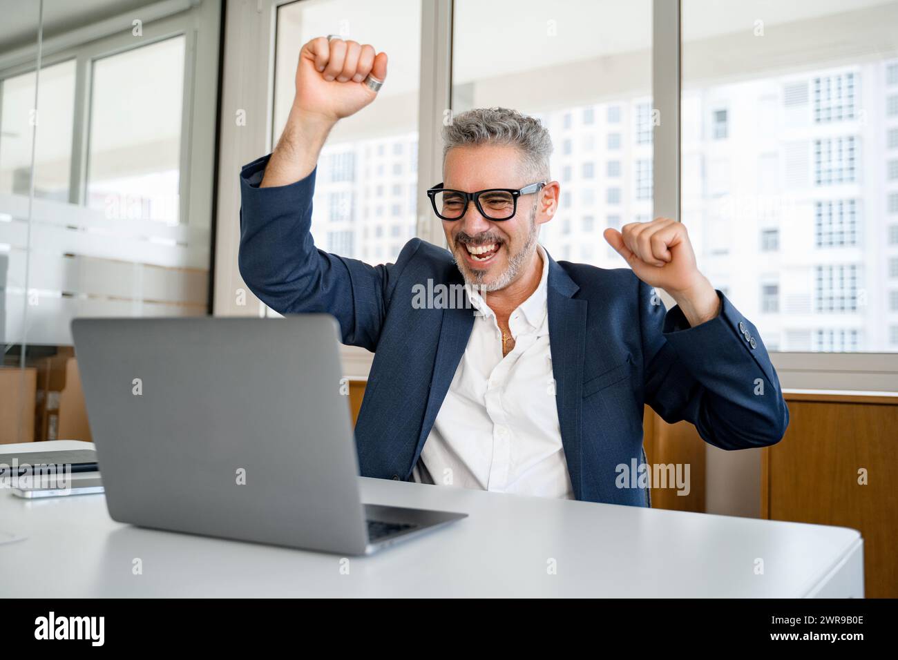 Overjoyed 60s mature businessman is gesturing victory with his arms raising up, senior manager scream with triumph, winning in game, having received great news of a good deal Stock Photo