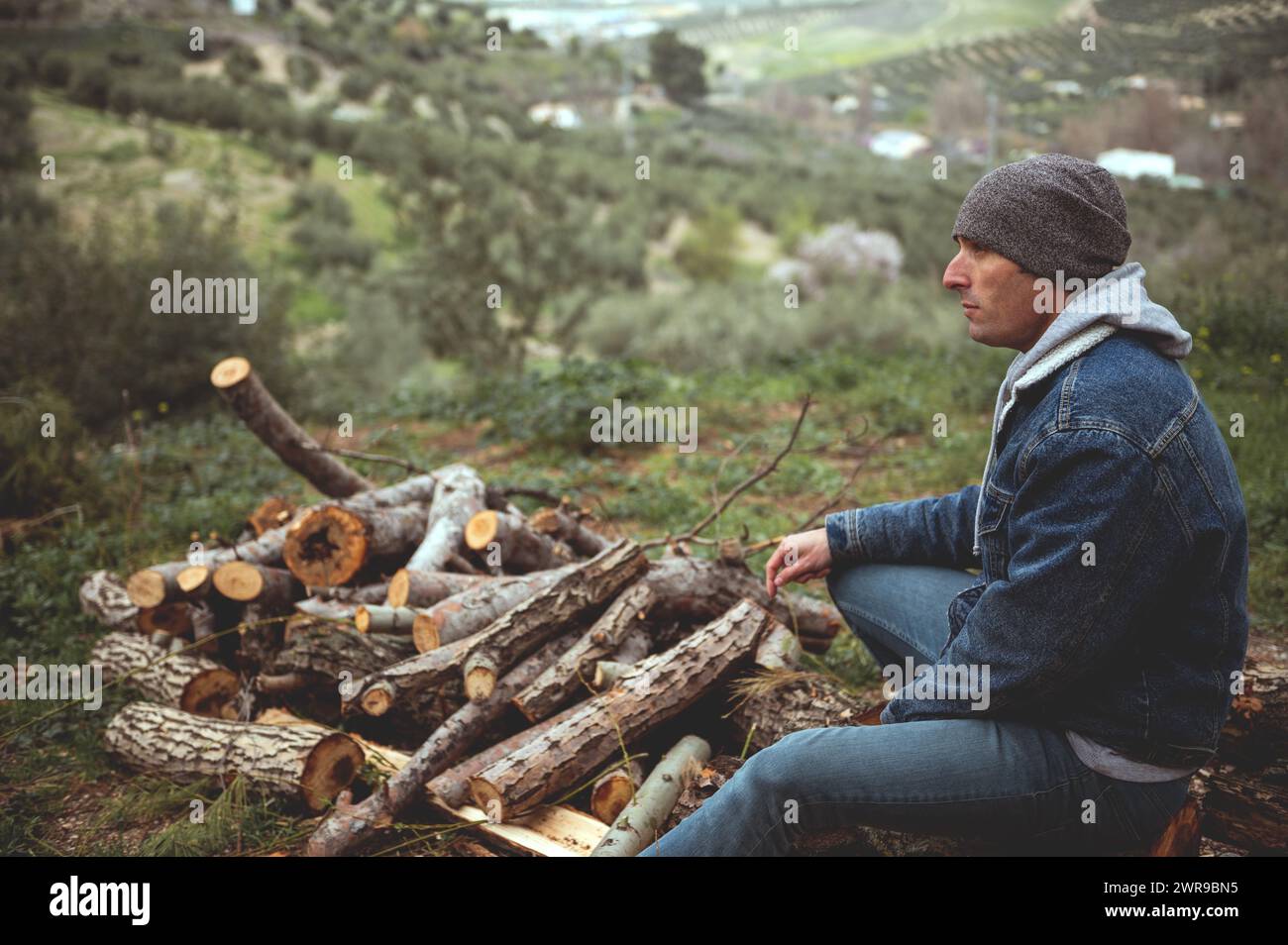 Handsome man sitting on a log near a pile of chopped firewood in forest. Young male hiker relaxing in the early spring nature, dressed in blue casual Stock Photo