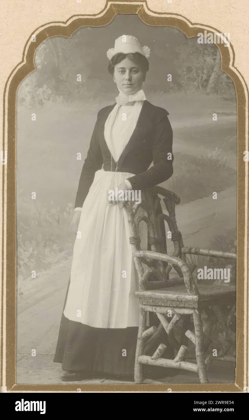 Portrait of a standing maid with a white hat, bow tie and apron by a chair. This photo is part of an album., E. v.d. Kerkhoff, Netherlands, 1887 - 1940, photographic support, gelatin silver print, height 85 mm × width 51 mm, photograph Stock Photo
