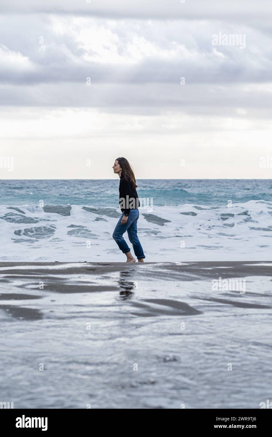 Adult woman with long brown hair walks alone and carefree on the shoreline barefoot. Full-length side view, casual wear with jeans and winter jumper. Stock Photo