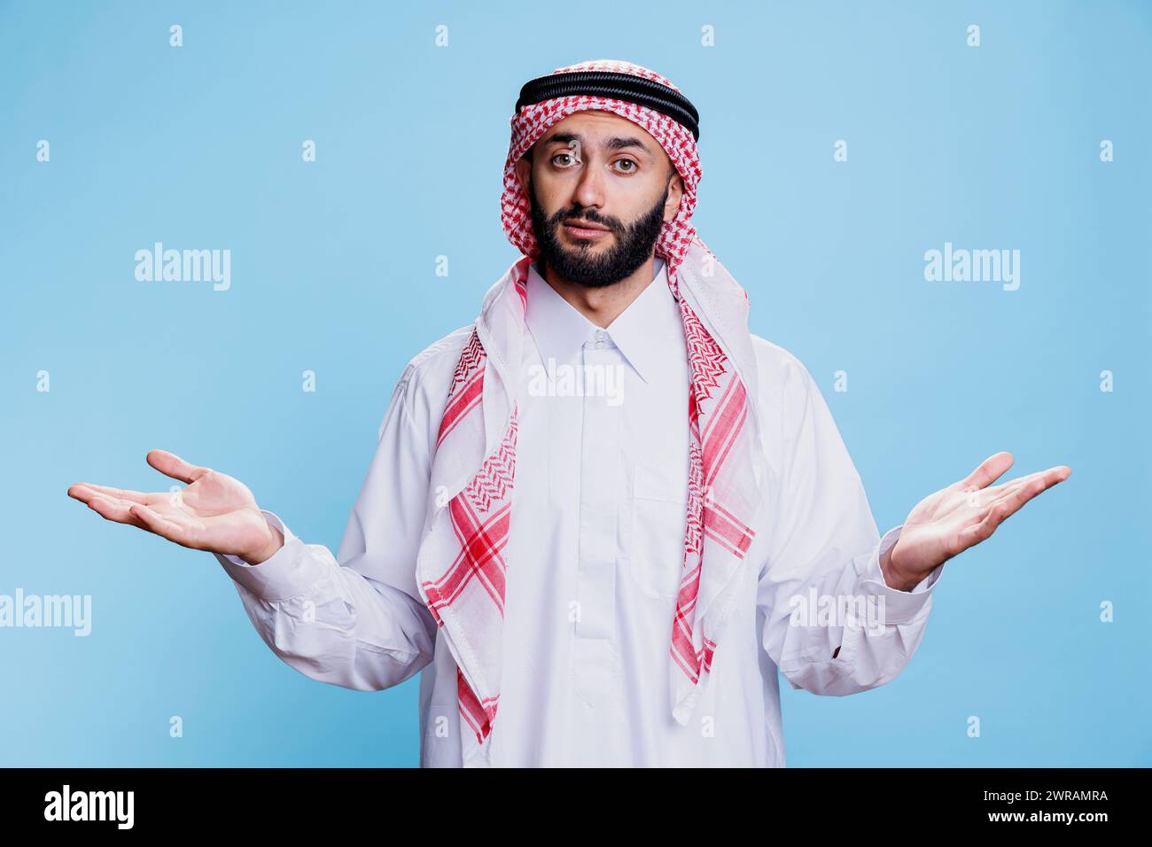 Pensive man wearing muslim thobe and headscarf, standing and shrugging shoulders while looking at camera. Arab person showing hesitation with open arms gesture studio portrait Stock Photo