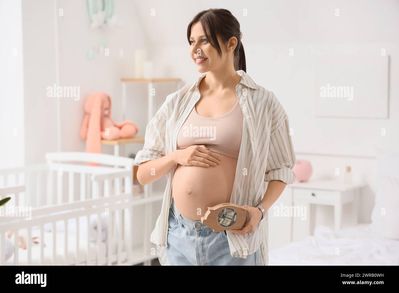 Young pregnant woman with piggy bank in bedroom. Maternal Benefit concept Stock Photo