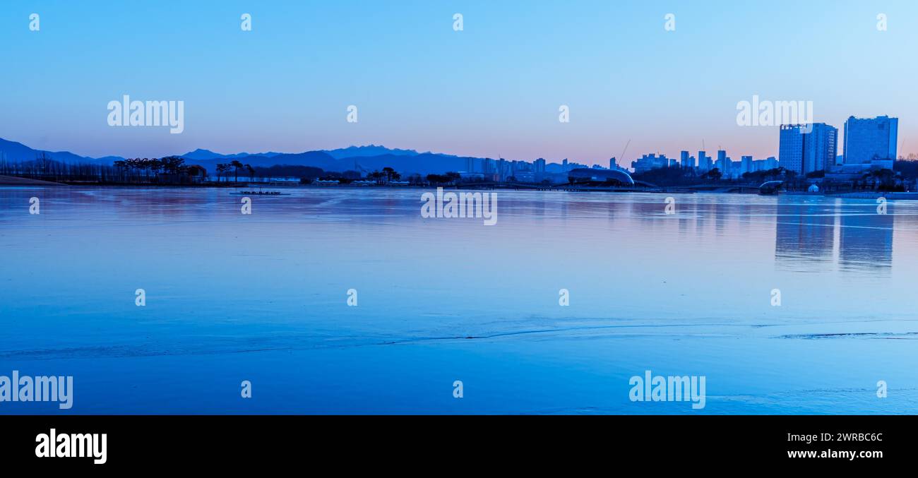 The city skyline reflects on calm waters at dawn, bathed in blue hues, in South Korea Stock Photo