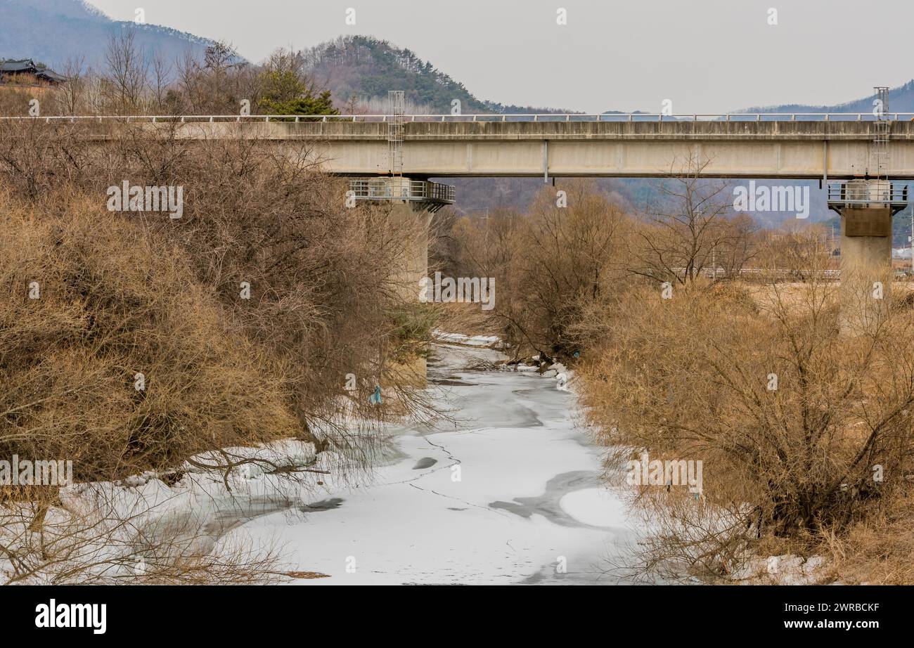 An overcast winter scene showing a bridge over a partially frozen creek with leafless trees, in South Korea Stock Photo