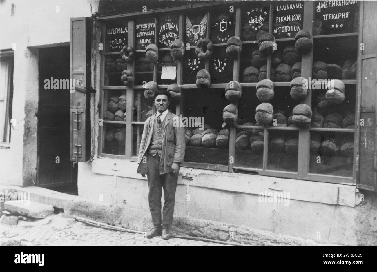Baker standing in front of the 'American Bakery' which displays signs in Armenian, Ladino (in Hebrew characters), English, Ottoman Turkish, Greek and Russian with samples of bread attached to the mullions, Ortakoy, Istanbul, Turkey, photo by cdm., Carpenter, Frank G. (Frank George), 1855-1924, collector, 1922 June., Bakeries, Turkey, Istanbul, 1910-1920, Photographic prints, 1910-1920., Portrait photographs, 1910-1920, Photographic prints, 1910-1920, 1 photographic print Stock Photo