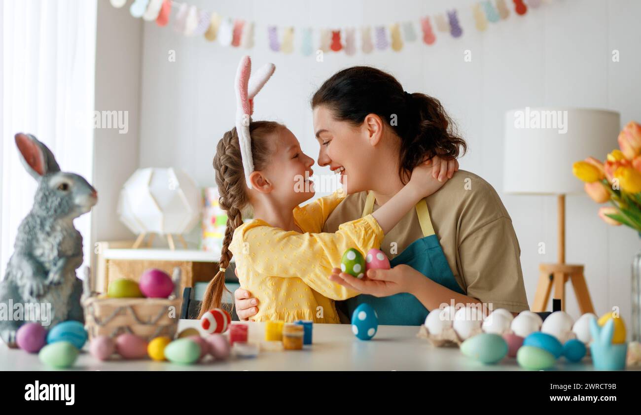 Mother and her daughter painting eggs. Happy family preparing for Easter. Cute little child girl wearing bunny ears. Stock Photo