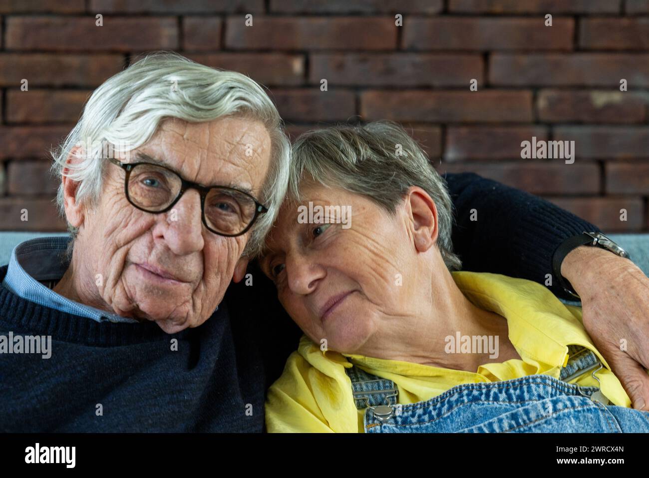 The image depicts an affectionate senior couple leaning on each other in a moment of relaxed intimacy. The man, with silver hair and glasses, shares the frame with a woman wearing a yellow top and denim overalls. Both appear content and comfortable with one another. The backdrop of a brick wall adds a rustic and homely feel to the scene, suggesting a setting of warmth and familiarity. Senior Couple Relaxed and Close Together. High quality photo Stock Photo