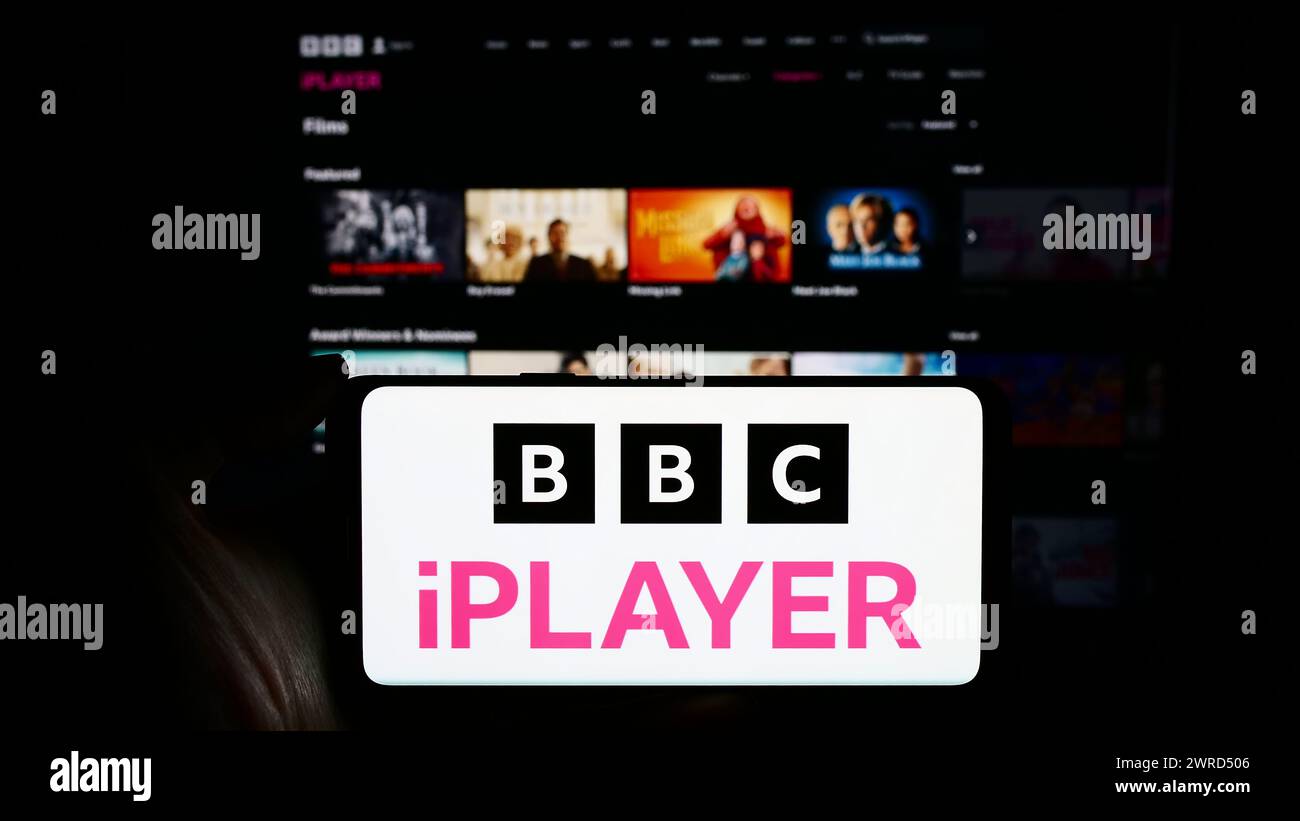 Person holding cellphone with logo of British video on demand service BBC iPlayer in front of business webpage. Focus on phone display. Stock Photo