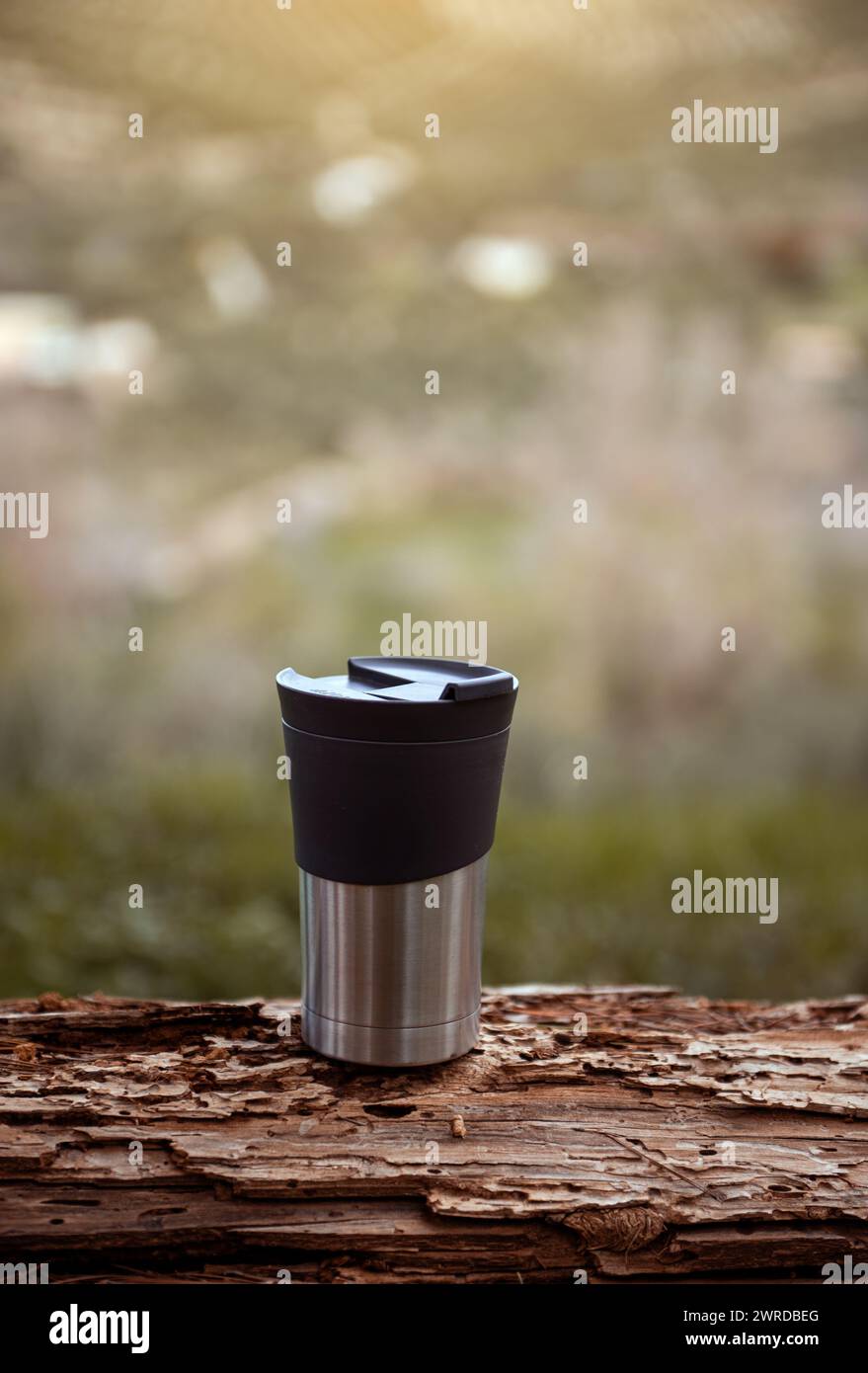 Vertical shot of a travel mug on a log over blur nature background with copy advertising space. A stainless steel cup with hot drink, coffee or tea fo Stock Photo