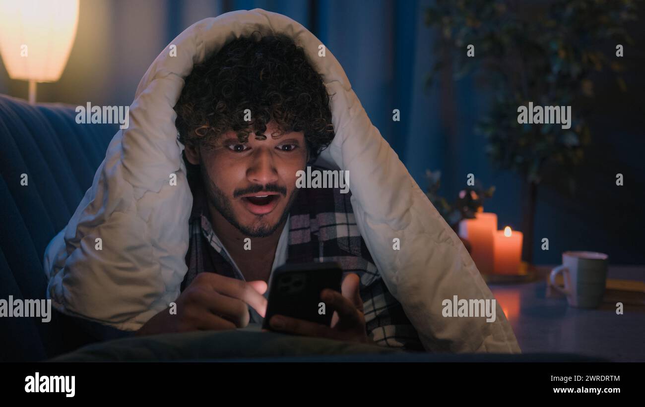 Mobile phone addict male user Indian guy surprised shocked wonder amazed reading news in smartphone at night under blanket Arabian man shock success Stock Photo