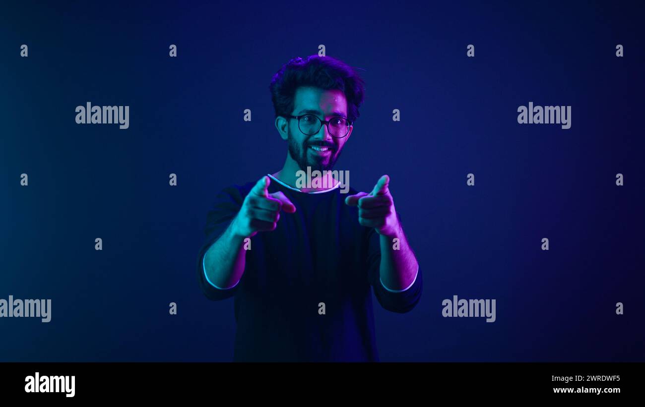 Portrait in neon ultraviolet happy Indian guy Arabian man worker coder hacker computer engineer make gesture hey you showing index finger front Stock Photo