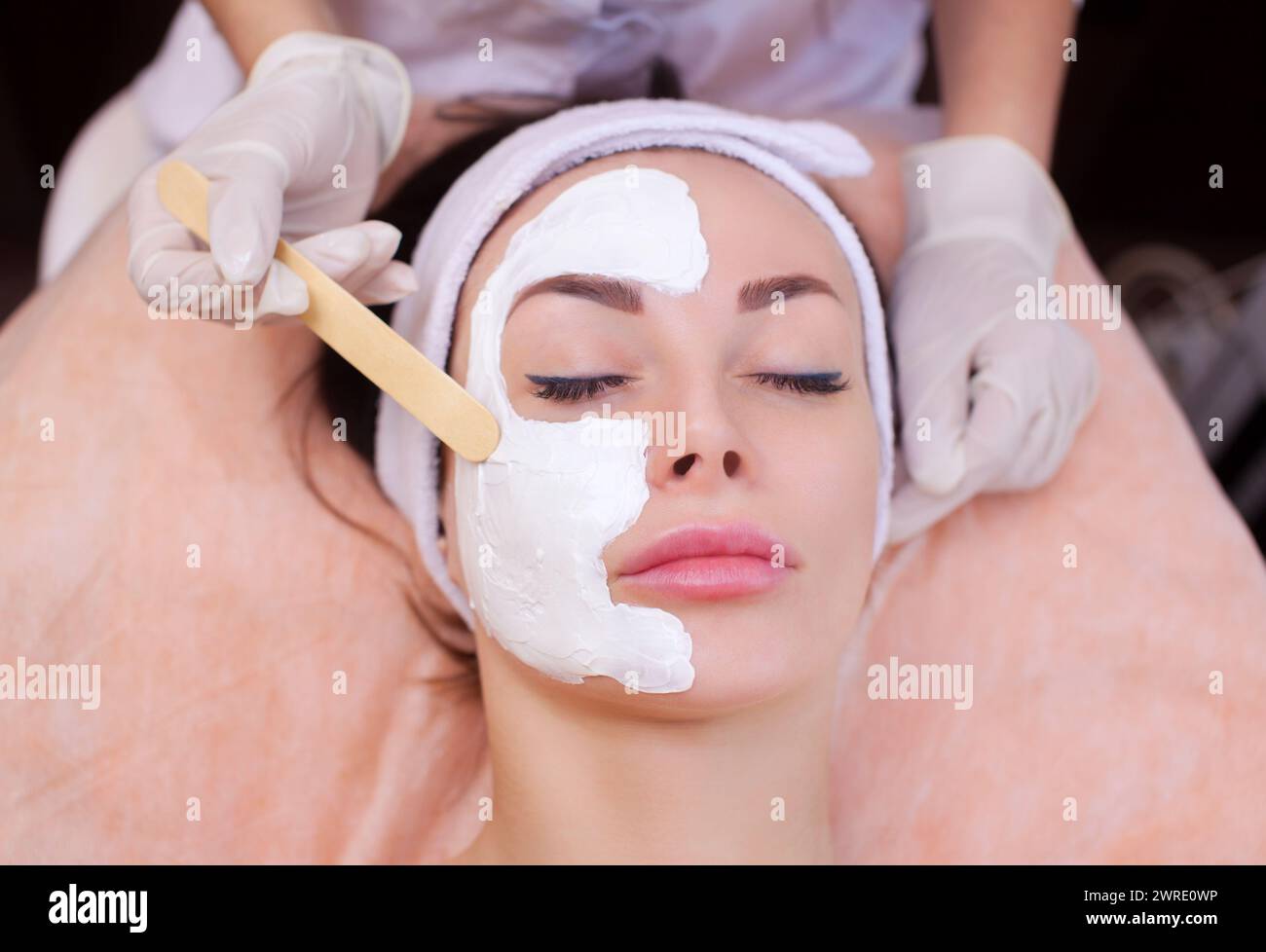 The doctor is a cosmetologist for the procedure of cleansing and moisturizing the skin, applying a mask with stick to the face of a young woman in bea Stock Photo