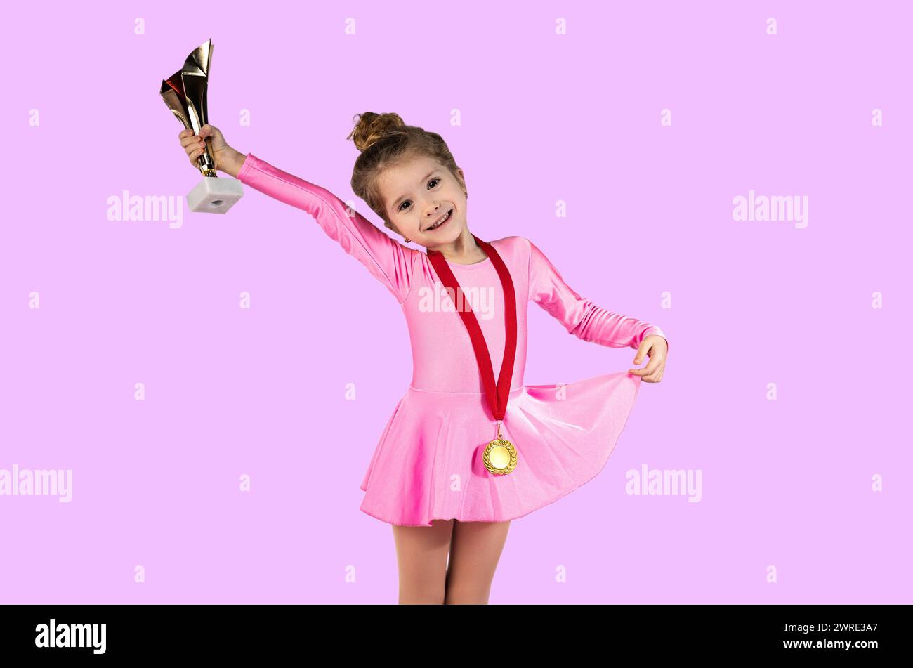 Portrait of a little gymnast girl right after winning dressed in a pink dress with a gold medal around her neck, Holding the newly won trophy in her h Stock Photo