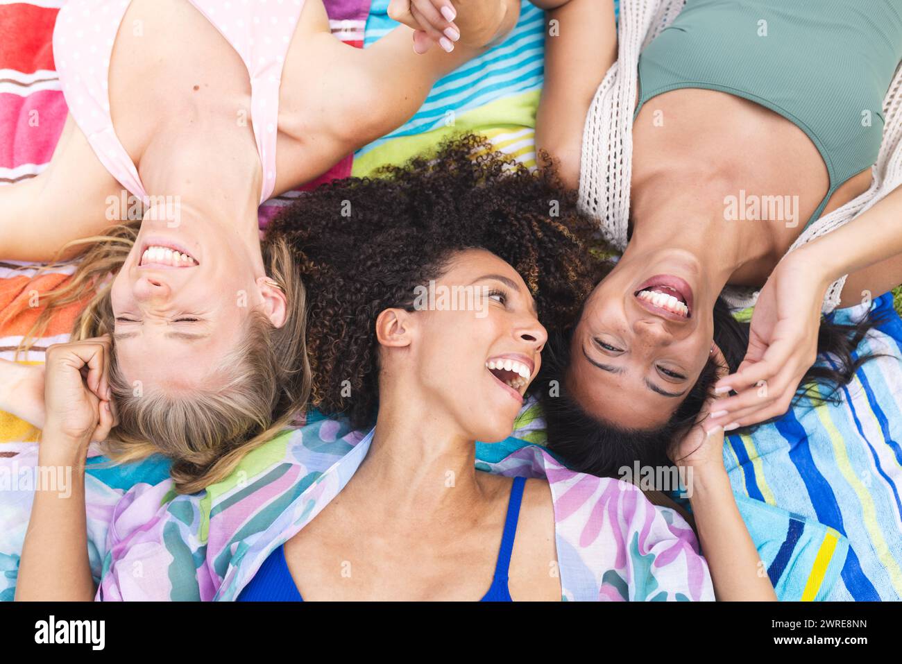 Diverse female friends are lying down, heads together, laughing joyfully Stock Photo