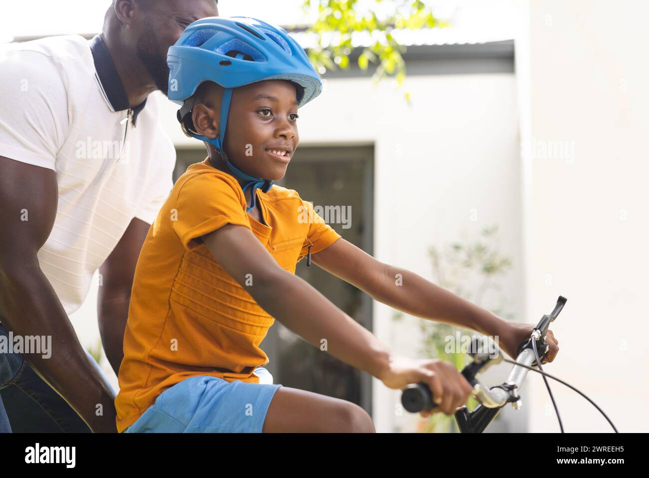 African American father teaches his son to ride a bike in the backyard, both smiling with joy Stock Photo