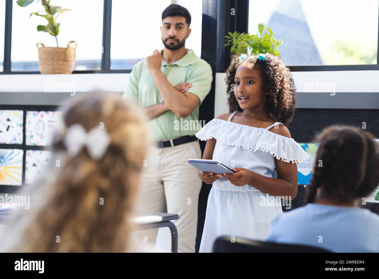 Young Asian male teacher stands observing a biracial girl presenting to the class in school Stock Photo