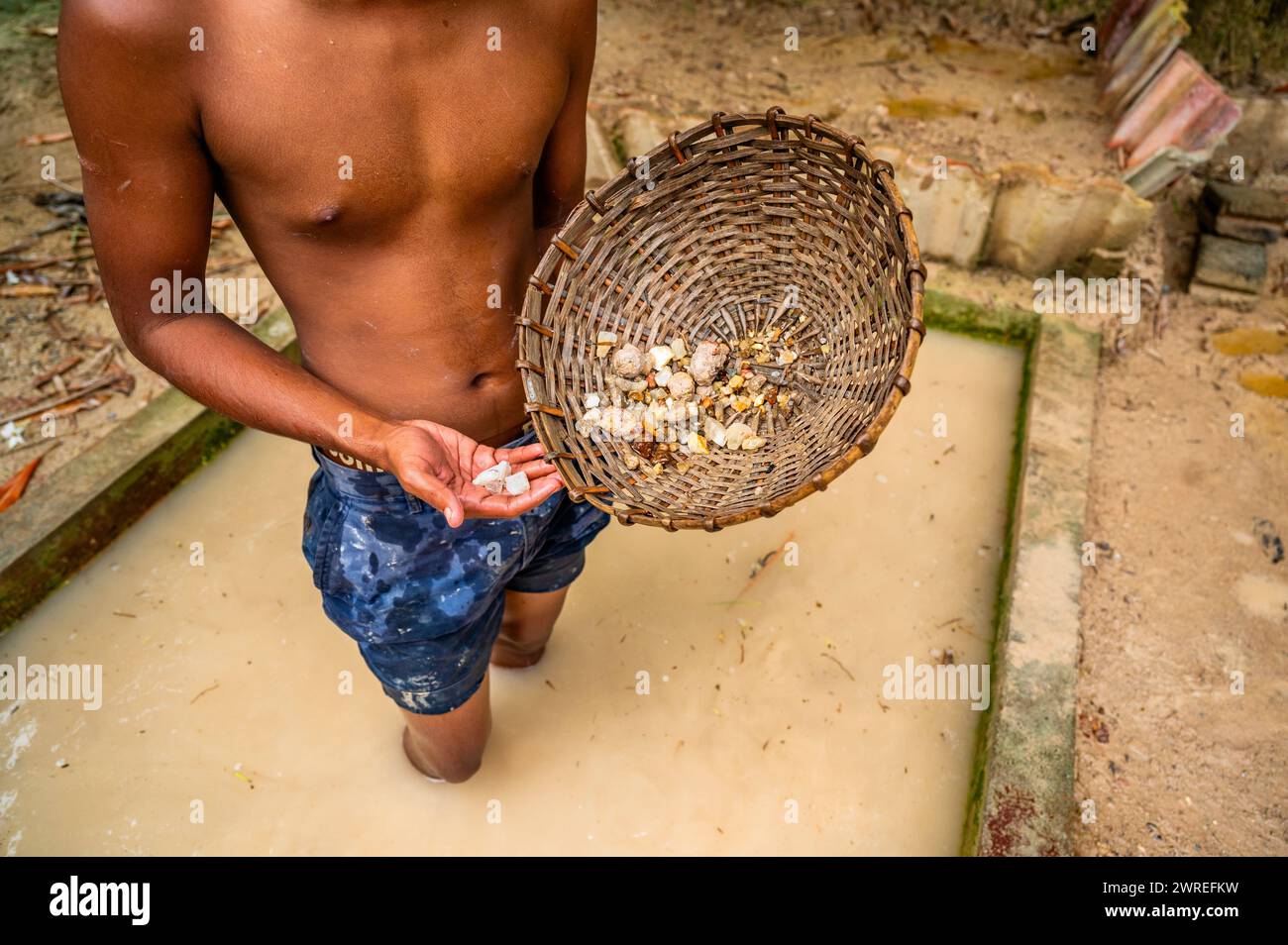 Boy in muddy water hold washed mined gemstone and rattan pan. Sri Lanka. Stock Photo