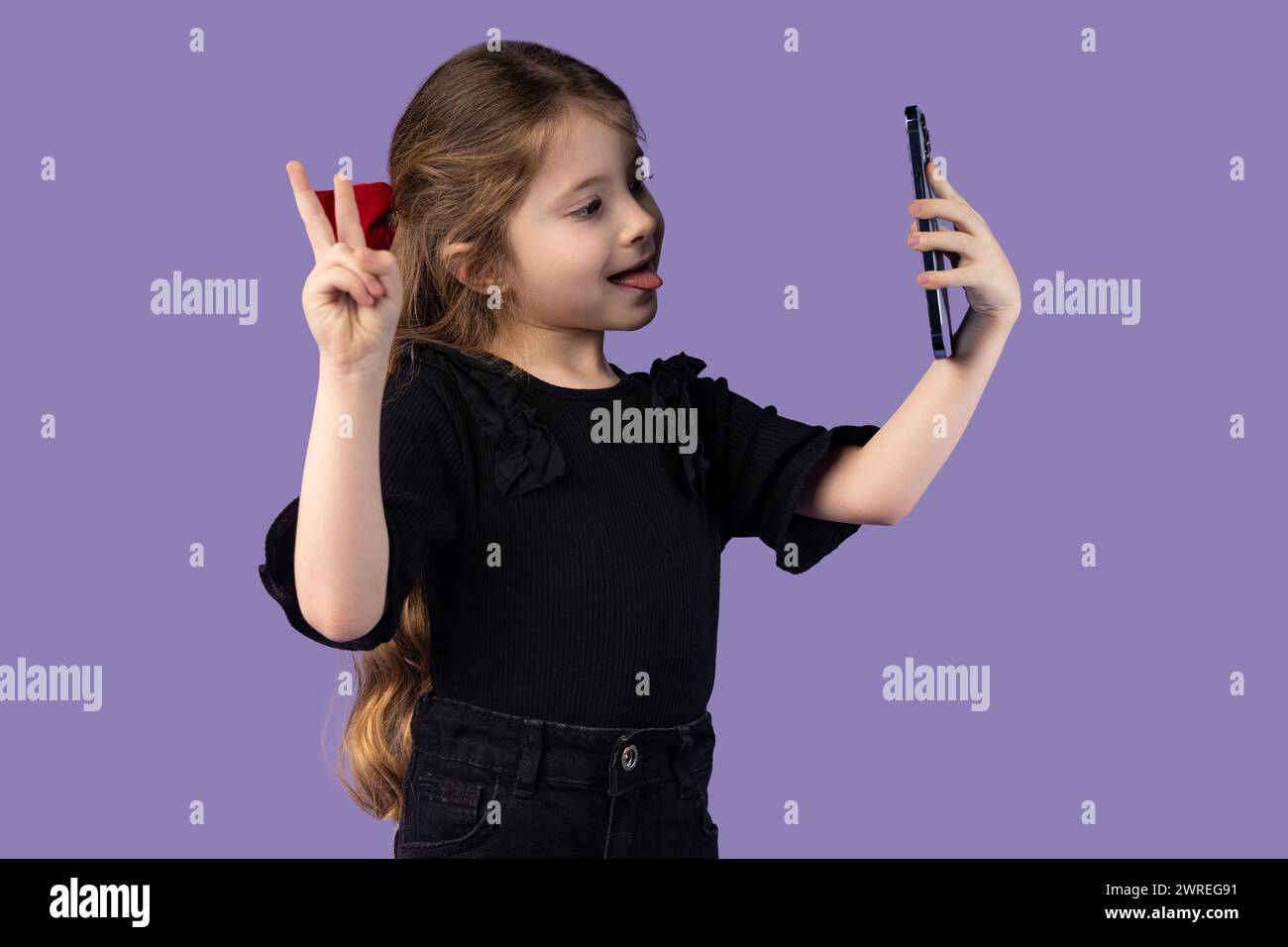 Photo of cute charming little girl in black t-shirt taking selfie with modern device showing isolated purple background with V sign. High quality phot Stock Photo