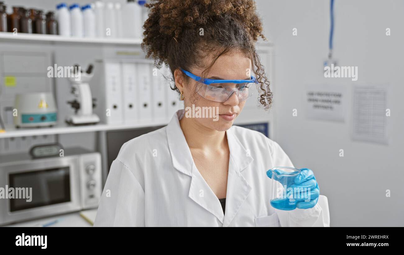 A young hispanic woman with curly hair wearing safety goggles and a lab ...
