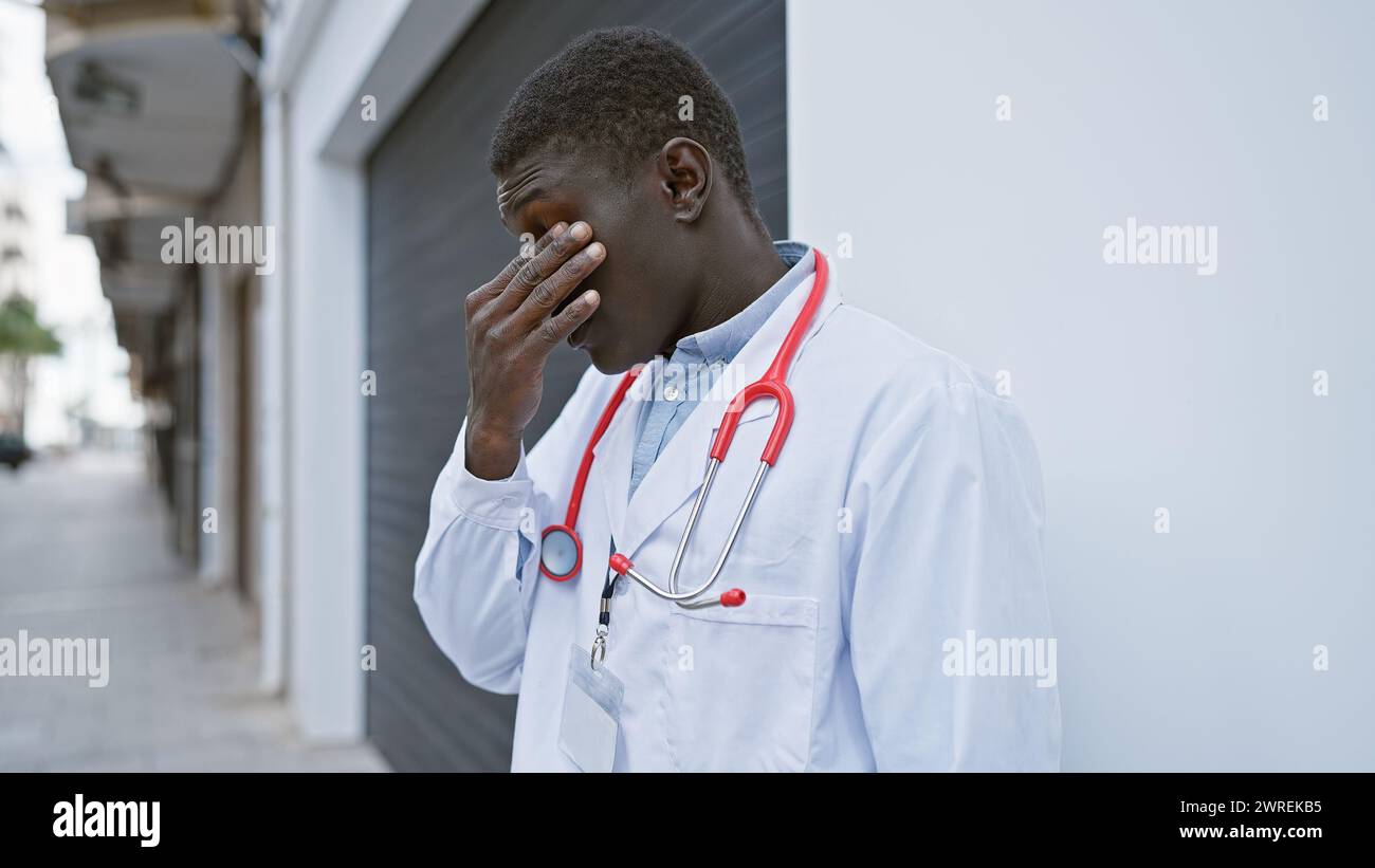 A tired black man in a doctor's coat with a stethoscope stands stressed on a city street. Stock Photo