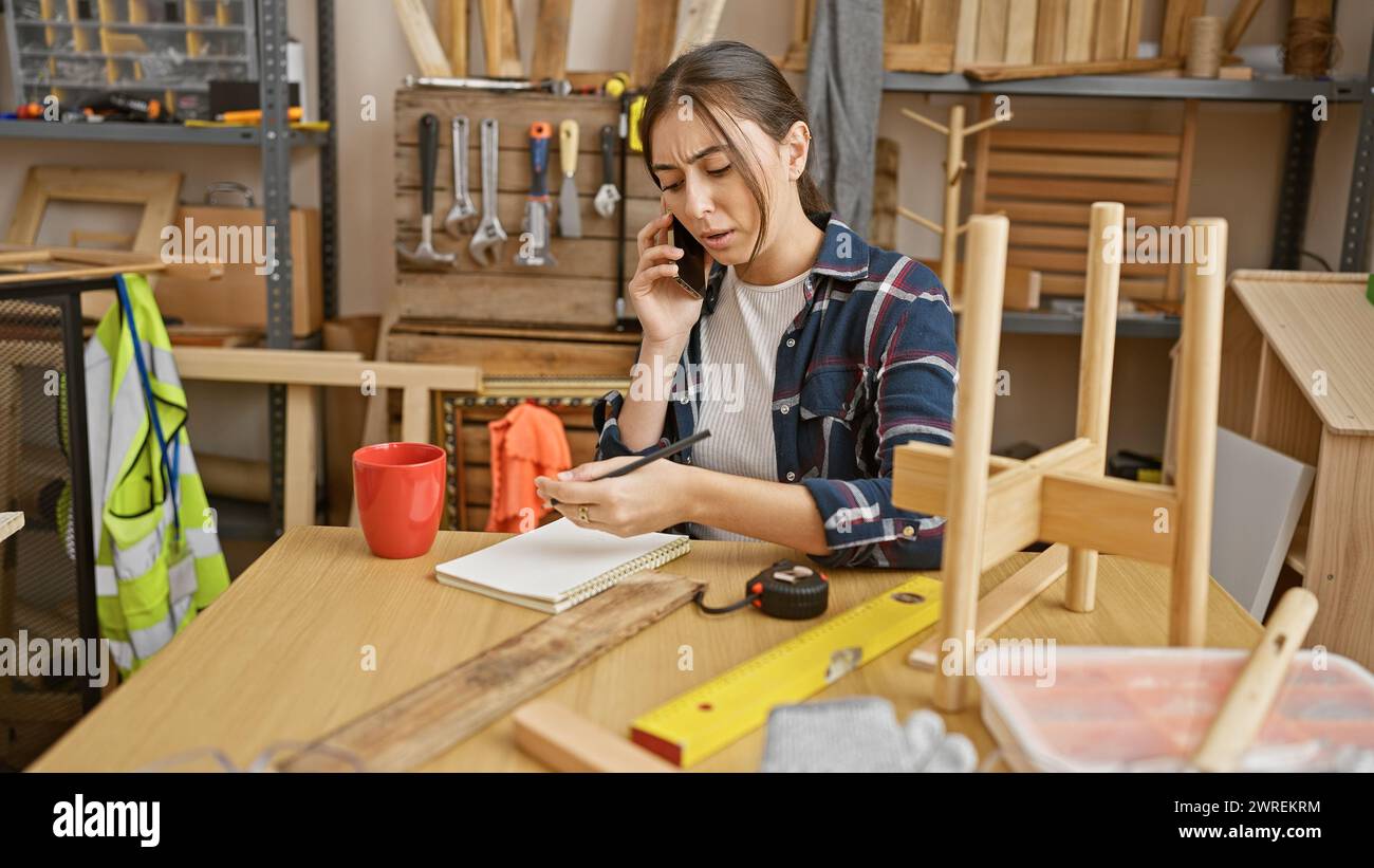 Hispanic woman multitasking in a carpentry workshop, talking on phone and taking notes. Stock Photo