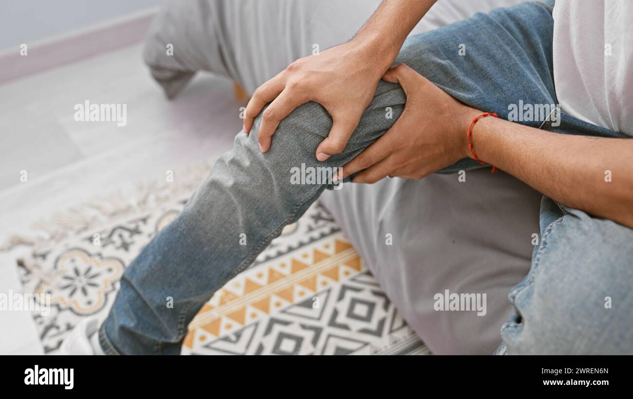 Young man holding knee in pain sitting on geometric-patterned rug at home, conveying a need for medical attention or injury recovery. Stock Photo