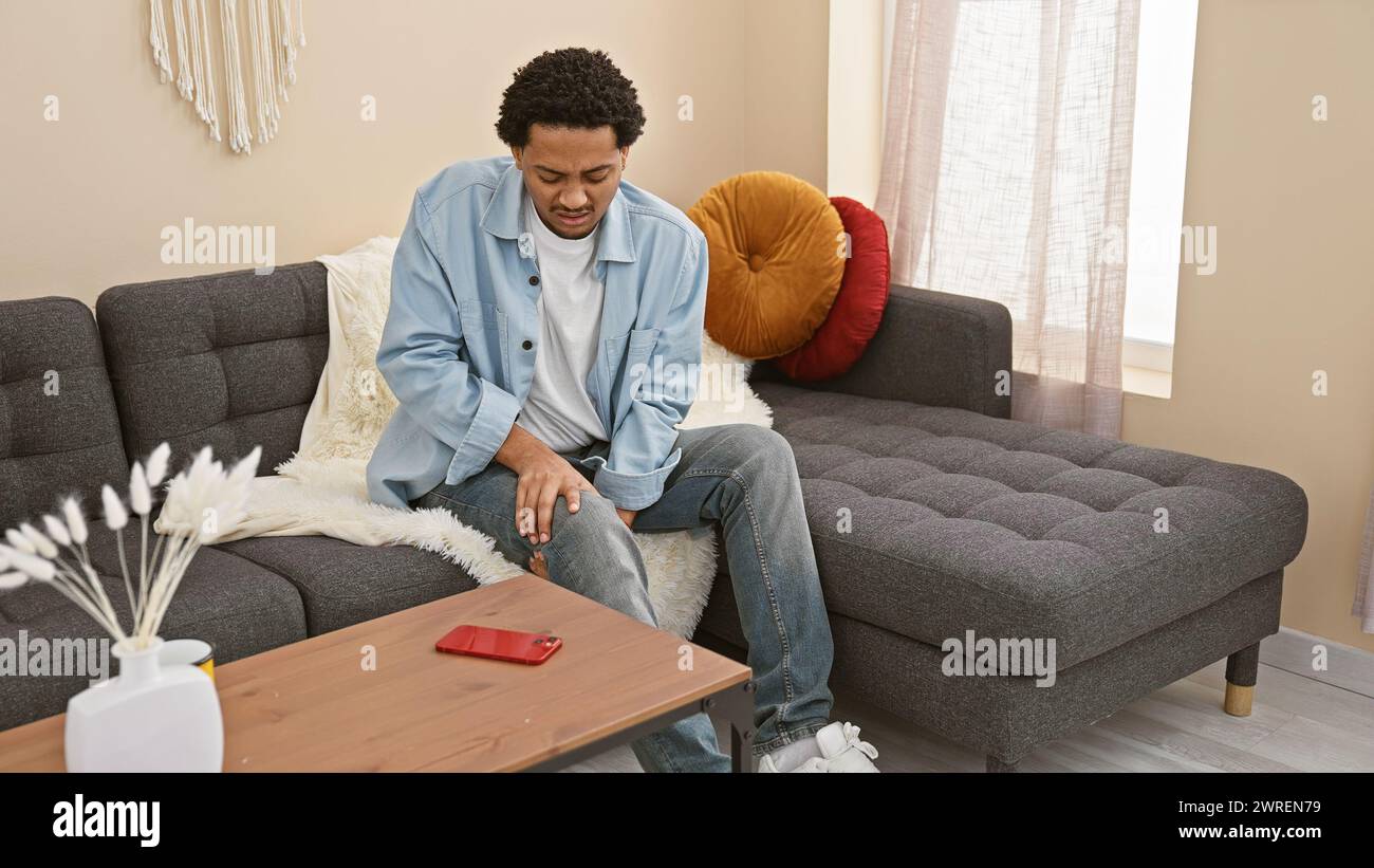 A young adult man with curly hair experiencing knee pain in a modern living room interior. Stock Photo