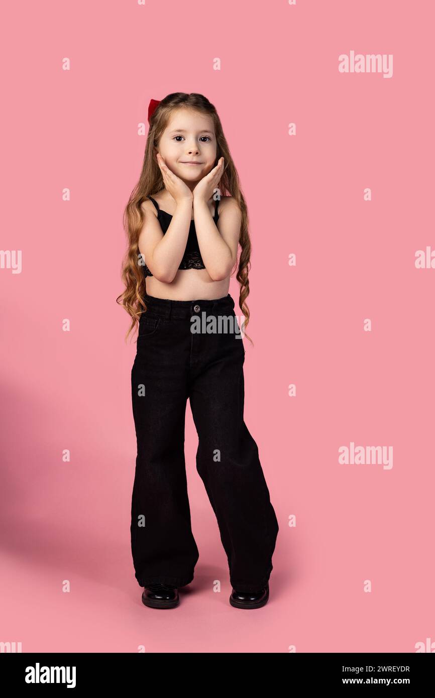 Full-length photo of a little girl standing with her hands under her chin, dressed in black looking at the camera photo taken on a pink background. Hi Stock Photo