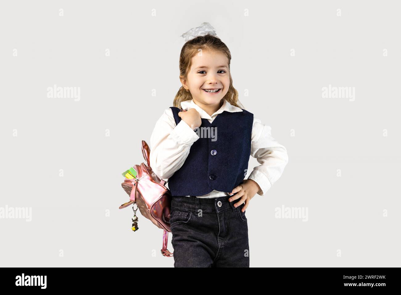 The little girl, with a smile on her face, is wearing a school uniform with puffed sleeves and carrying a backpack. She looks happy and her thumbs are Stock Photo